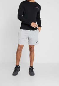 Černá tílko s dlouhými rukávy a malým logem, doplněné o světle šedé kraťasy a černé sportovní boty. Tkanina vypadá hladce a je lehká.