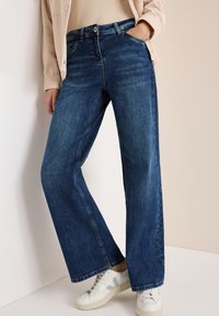 Wijdvallende blauwe denim jeans met een middelhoge taille, vijf zakken en subtiele vervaging. Gemaakt met een lichtgekleurde sneaker.