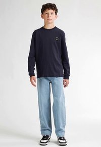 Námořnicky modrá košile s dlouhým rukávem a kulatým emblémem, doplněná světle modrými rovnými džínami a černými teniskami s bílými detaily.