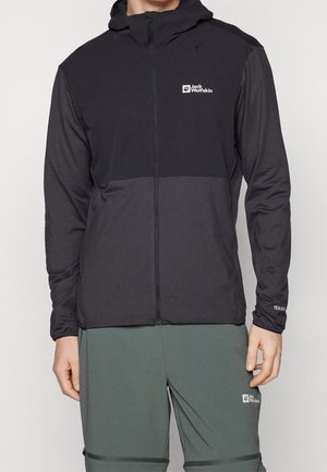 Man wearing black and dark grey Jack Wolfskin hooded jacket and green Jack Wolfskin pants, standing against a plain background.