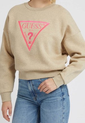 Beżowa bluza z różowym trójkątem z logo Guess i niebieskie dżinsy noszone przez osobę z ręką opartą na biodrze.