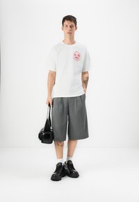 T-shirt blanche avec un graphique rouge, short plissé gris, baskets noires et un petit sac noir. Design simple et coupe décontractée.