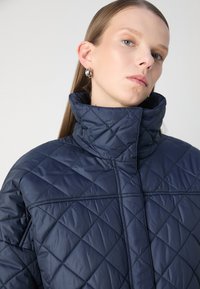 Μπλέ πλισέ μπουφάν με ψηλό γιακά, με διαχρονικά μοτίβα διαμαντιού και απαλή, γυαλιστερή υφή. Κομψός, μινιμαλιστικός σχεδιασμός.