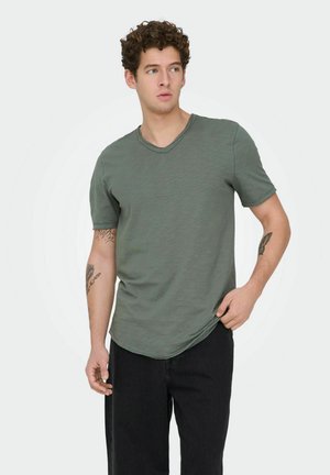 Young man with curly hair wearing a green V-neck T-shirt and black pants, standing and looking to his left with tattoos visible on both arms.