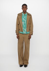 Brown trench coat over a green striped shirt with floral accents, paired with beige wide-leg trousers and black shoes. Plain white background.