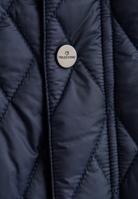 Marineblauer, gesteppter Stoff mit glänzendem Finish, verziert mit Rautenmuster, gesteppten Details und einem Metall-Druckknopf mit dem Logo "Milestone" geprägt.