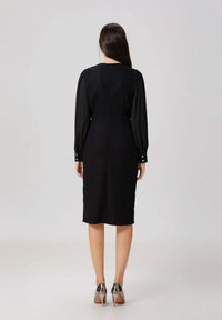 Robe noire à manches longues, taille cintrée et ourlet à hauteur du genou. Présente une texture subtile et une fermeture éclair au dos. Portée avec des talons hauts métalliques.