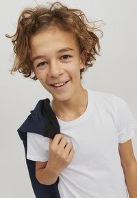 T-shirt bianco a maniche corte indossato da un ragazzo giovane, che tiene un felpa blu scuro sulle spalle, con capelli ricci e uno sfondo neutro.