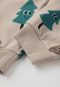 Beige sweatshirtmouw met groene boom- en dennenappelprint. Geribbelde boorden, zacht katoenen materiaal, speelse ontwerpdetails met glimlachende elementen.
