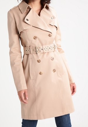 Beiger, zweireihiger Trenchcoat mit weitem Kragen, tailliertem Gürtel und seitlichen Taschen, getragen über dunkelblaue Jeans.