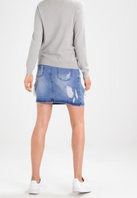 Personne portant un haut à manches longues gris clair, une jupe en denim bleu déchiré et des baskets blanches, debout de dos sur un fond blanc uni.