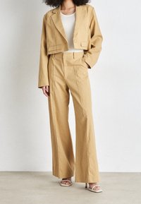 Beżowa krótka marynarka zestawiona z szerokimi spodniami, oba elementy charakteryzują się czystymi liniami i widocznym przeszyciem. Modelka ma na sobie sandały na obcasie na neutralnym tle.