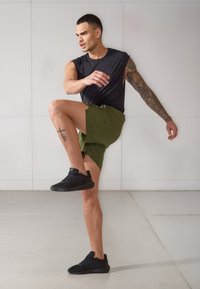 Μαύρο αμάνικο αθλητικό μπλουζάκι, ελιά χρώμα σορτσάκι και μαύρα αθλητικά παπούτσια. Δείχνοντας ανύψωση ποδιού; αξιοσημείωτο τατουάζ στη γάμπα. Λείο, μοντέρνο περιβάλλον.