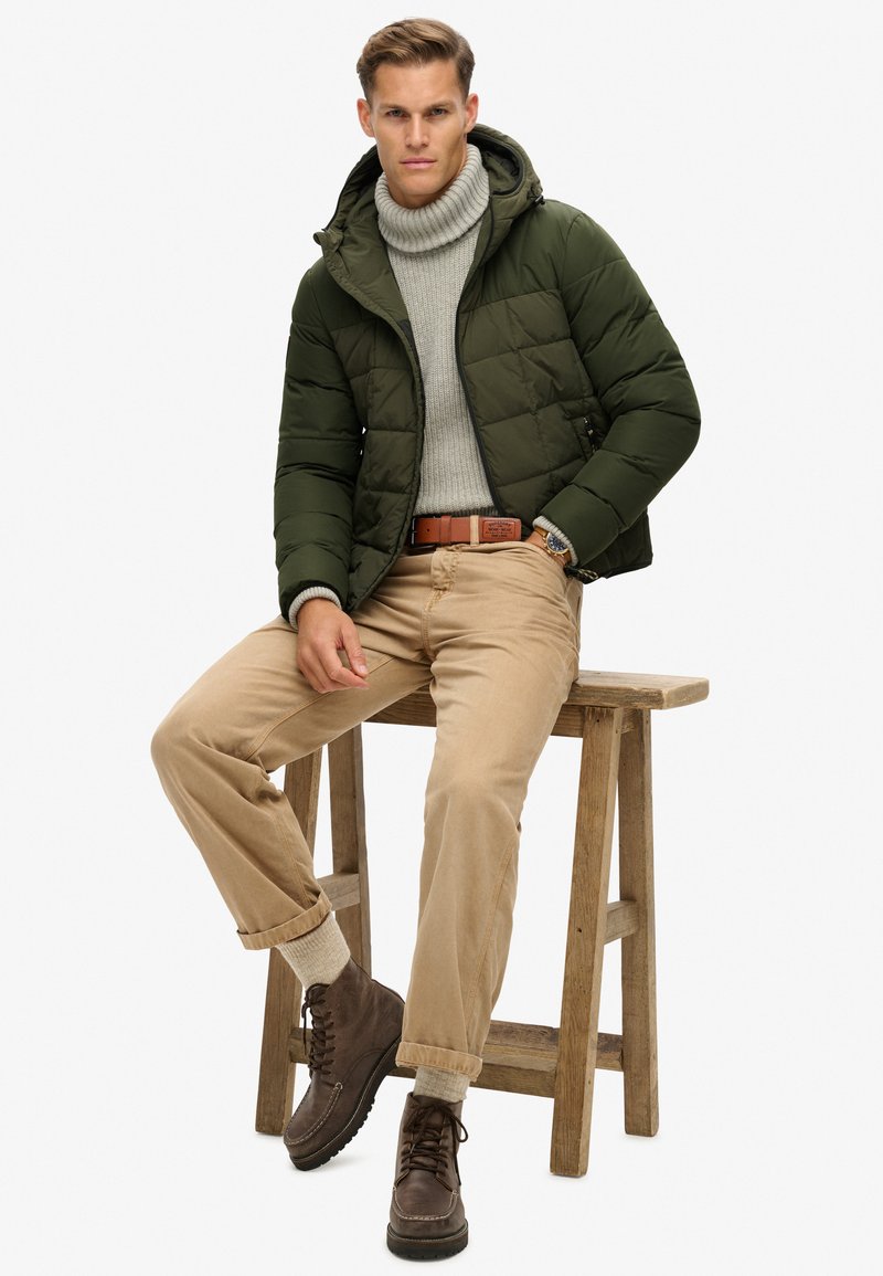 Giacca imbottita verde con cappuccio, dolcevita grigio, chinos beige, stivali marroni, seduto su uno sgabello di legno. Tessuti strutturati e design sobrio.