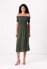 Off-the-shoulder, smockad klänning i olivgrön färg med flowy kjol, knälång och lätt material, matchad med beige remsandaler.