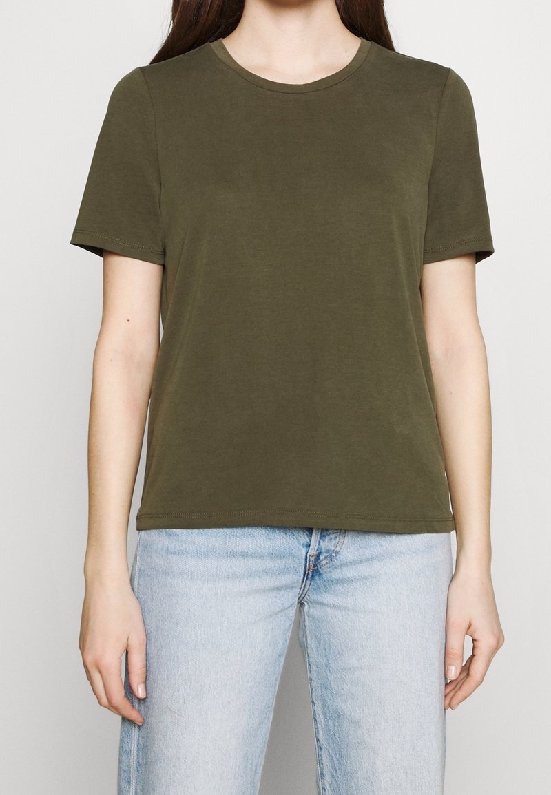 Femme portant un t-shirt uni vert olive à manches courtes et col rond, ainsi qu'un jean bleu clair, debout devant un fond uni.