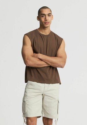 Giovane uomo con capelli corti in piedi con le braccia incrociate, indossa una maglietta marrone senza maniche e pantaloncini cargo beige su uno sfondo semplice.