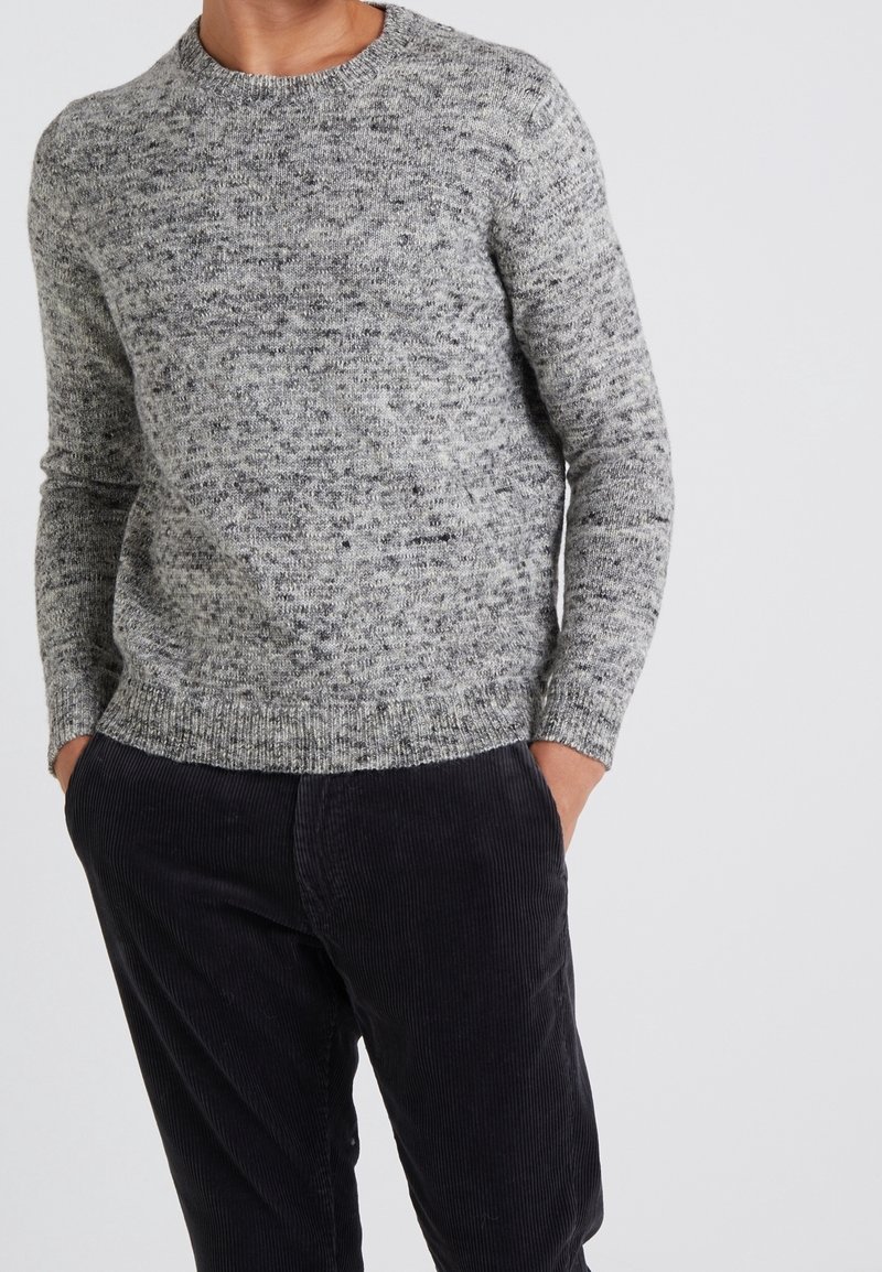 Pull en tricot gris avec une finition texturée, encolure ronde et poignets côtelés. Porté avec un pantalon en velours côtelé foncé.