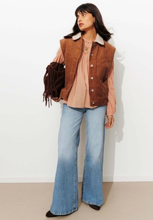 Gilet sans manches en velours côtelé marron avec un col en fausse fourrure, superposé sur un haut rose clair, associé à un jean bleu clair évasé et un sac à franges marron.