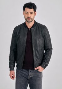 Man met zwart haar die een zwart leren jack, een donkere trui en grijze jeans draagt, staand met één hand in zijn zak.