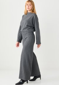 Frau steht, trägt ein passendes graues langärmeliges Oberteil und weit geschnittene Hosen mit schwarzen, hochhackigen Stiefeletten vor weißem Hintergrund.