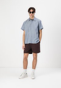 Kurzarmhemd aus leichtem Blau mit vertikalen Streifen, kombiniert mit dunkelbraunen Shorts und weißen Sneakern, ergänzt durch schwarze Sonnenbrillen.
