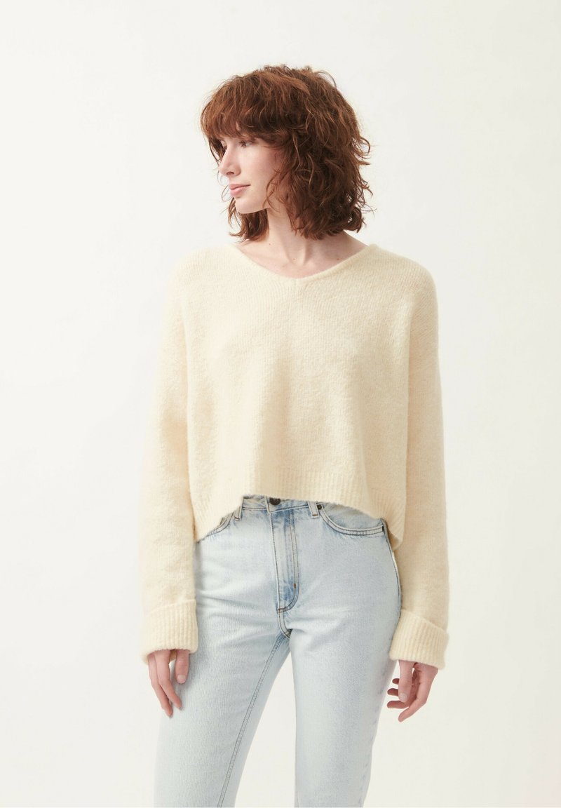 Pull cropped couleur crème avec un col en V, des poignets côtelés et une texture douce, associé à un jean bleu clair.