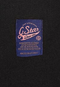 Tessuto nero con un motivo testurizzato che presenta un'etichetta blu. L'etichetta mostra "G. Star Raw" in scrittura rosa con istruzioni per la cura.