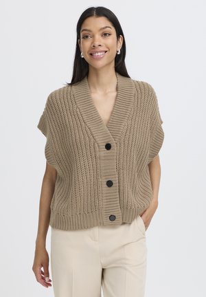 Femeie zâmbitoare purtând un cardigan bej gros fără mâneci, cu nasturi negri, și pantaloni crem deschis, cu mâna în buzunar, pe fundal alb.