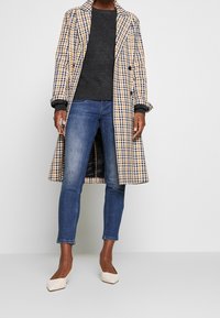 Abrigo de lana a cuadros en azul, amarillo y blanco; suéter gris; jeans azules; zapatos planos de punta beige. Presenta un diseño de doble botonadura.
