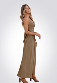 Tutina beige senza maniche con un corsetto aderente e gambe ampie e fluide. Indossata con scarpe con tacco aperte di colore marrone e mentre tiene una borsa piccola.