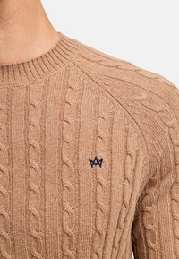 Kabelstrikket genser i lys brun med ribbet krage. Har teksturert mønster og en marineblå logo aksent på brystet.