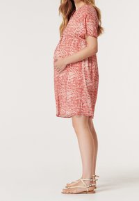 Röd mönstrad gravidklänning med korta ärmar och lös passform som framhäver en rundad mage. Bärs med beiga remsandaler.