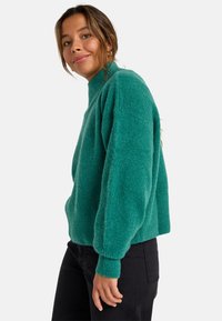 Turkusowy, oversize sweter z wysokim kołnierzem i prążkowanymi mankietami, wykonany z miękkiego, teksturowanego materiału. Połączony z czarnymi spodniami.