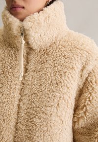 Veste en polaire beige à texture bouclée, avec un col haut et une demi-fermeture éclair, portée par une personne avec de petites boucles d'oreilles et un maquillage des lèvres aux tons neutres.