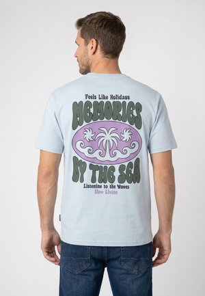 Man facing away wears light blue t-shirt with palm tree design and text "Feels Like Holidays Memories By The Sea Listen to the Waves Slow Living."