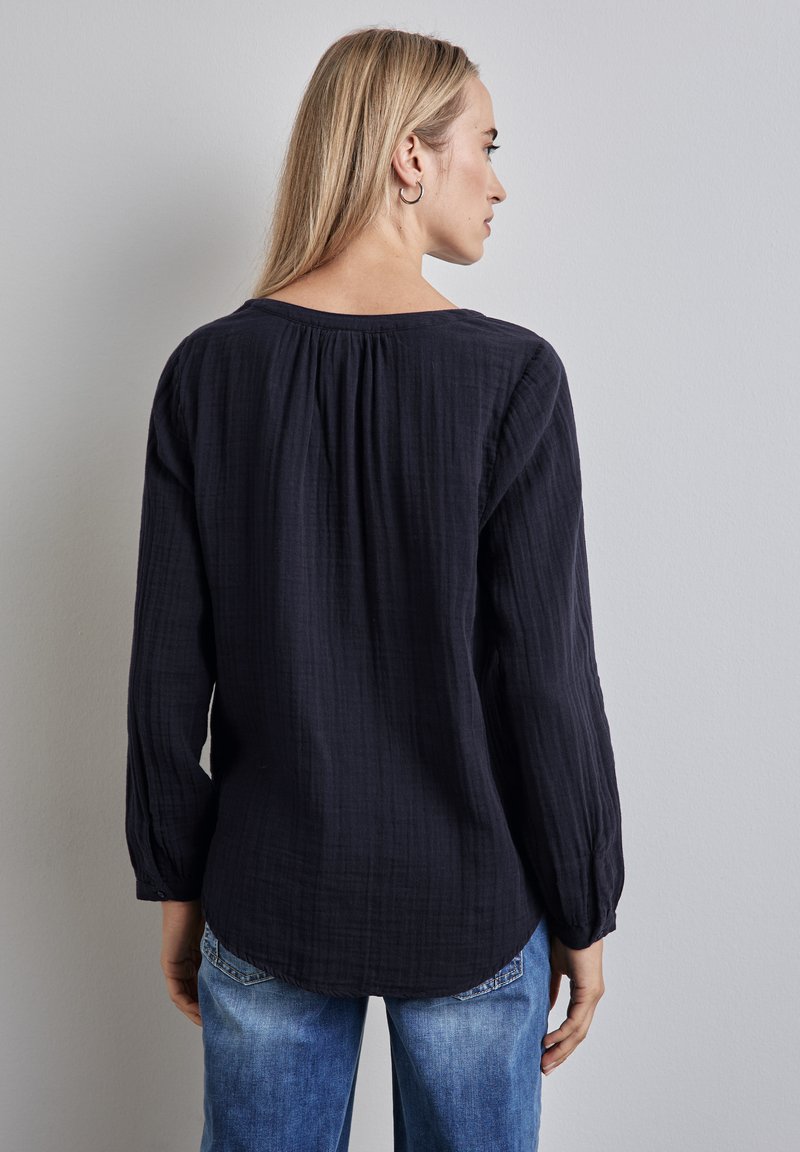 Maglietta a maniche lunghe di un blu navy scuro con tessuto texture, dotata di scollatura rotonda e dettaglio arricciato sul retro, abbinata a jeans blu chiaro.