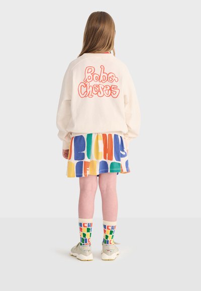 Bambino con felpa crema con la scritta rossa "Bobo Choses" sulla schiena, gonna colorata con motivo, calzini abbinati e sneakers chiare, in piedi di spalle.