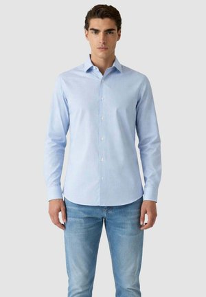 Giovane uomo in piedi, rivolto in avanti, che indossa una camicia a righe azzurre con bottoni e jeans blu, su uno sfondo semplice.