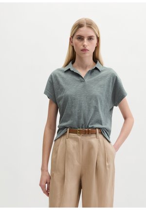 Femme aux longs cheveux blonds porte une chemise à manches courtes col boutonné bleu ardoise, rentrée dans un pantalon plissé beige avec une ceinture marron.