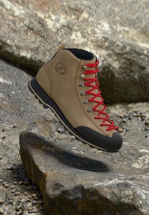 Au-dessus d’un sentier rocailleux, on voit une chaussure de randonnée beige, avec des lacets rouge vif et une semelle noire.