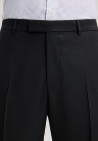 Schwarze Hose aus strukturiertem Stoff, mit flacher Vorderseite, Gürtelschlaufen und einem eleganten Design, kombiniert mit einem weißen Hemd mit Polka-Dots.