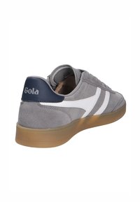 Sneaker in suede grigio con strisce bianche e un accento blu sul tallone, dotato di suola in gomma e lacci piatti. Logo del marchio visibile sul tallone.