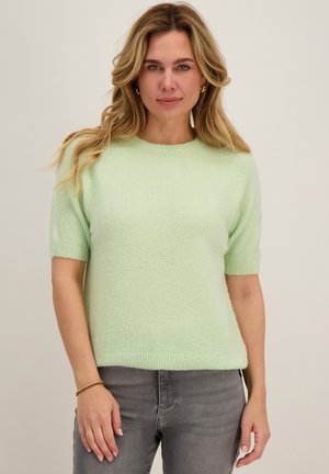 Vrouw met golvend blond haar, met gouden creolen en een muntgroene trui met korte mouwen, gecombineerd met grijze spijkerbroek, staand tegen een effen achtergrond.