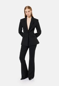 Sort skræddersyet blazer med spidse klapper og en enkelt knaplukning, parret med matchende tætsiddende bukser. Modellen står med hænderne på hoften.