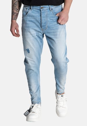 Light blue denim jeans with a faded design, featuring a ripped detail on the left knee, and five pockets. Worn with white sneakers.