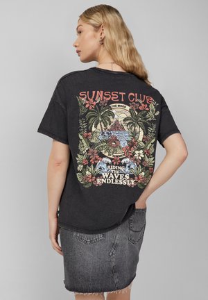 Black oversized t-shirt featuring a vibrant tropical graphic with flowers, palm trees, and text on the back. Combined with a gray denim skirt.