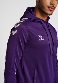 Felpa con cappuccio viola caratterizzata da una texture liscia, logo bianco e dettagli sulle spalle, con tasca frontale e lacci regolabili.