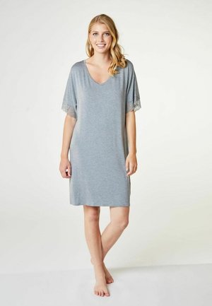 Donna sorridente con lunghi capelli biondi, in piedi a piedi nudi, indossa un abito largo grigio chiaro a maniche corte con bordo in pizzo sulle maniche.