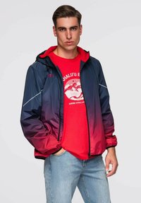 Mladý muž v červeném tričku s grafikou a modro-červené přechodové větrovce s kapucí, stojící s jednou rukou v kapse.